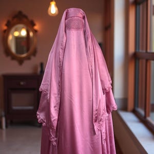 Può includere: Viene mostrato un burqa rosa, un indumento tradizionale. Il burqa è realizzato in un tessuto liscio, simile al raso, e presenta intricati dettagli in pizzo lungo i bordi. Il design include un copricapo e un velo che copre il viso.