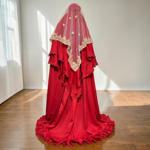 Può includere: Un abito in raso rosso con gonna a strati e orlo a balze. L'abito è abbinato a un velo trasparente ornato di pizzo dorato e decorazioni floreali. Adatto per un'occasione speciale.