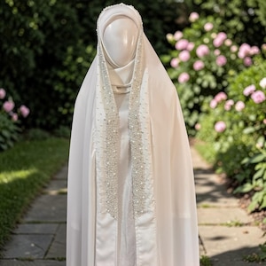 May include: A white garment with a head covering and a long, flowing cape. The garment is adorned with rows of small, white beads along the front edges. The background features a stone path and a garden with pink flowers.