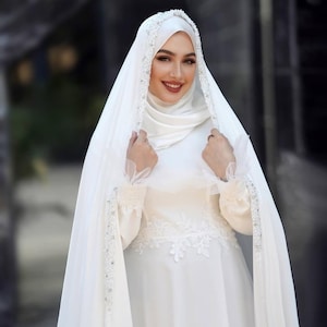 May include: A white wedding dress with a matching head covering and veil. The dress features delicate lace detailing and is adorned with sparkling embellishments. The veil is draped over the shoulders, creating an elegant and modest look.