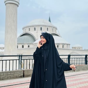 Op de afbeelding: Een vrouw draagt een zwarte abaya met een lange, vloeiende rok en een bijpassende hoofddoek. Ze staat voor een moskee met een witte koepel en minaretten.