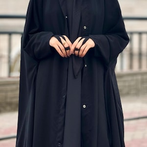 Op de afbeelding: Een zwarte abaya met een lang, vloeiend ontwerp en een knoopsluiting. De abaya heeft een capuchon en lange mouwen.