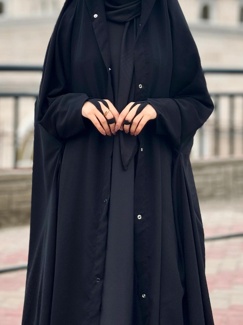 Op de afbeelding: Een zwarte abaya met een lang, vloeiend ontwerp en een knoopsluiting. De abaya heeft een capuchon en lange mouwen.
