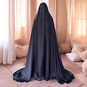 Puede incluir: Una prenda de satén azul marino oscuro, que cubre todo el cuerpo, con una cola fluida. La tela se drapea elegantemente, creando pliegues y sombras suaves. La figura está de pie en una habitación con iluminación suave y cojines decorativos.