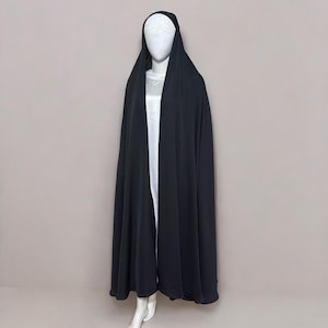 Peut inclure: Un abaya noir, un long vêtement fluide, est drapé sur un mannequin. L'abaya est doté d'une capuche et couvre le mannequin de la tête aux pieds. Un vêtement blanc est visible en dessous. L'arrière-plan est de couleur neutre.