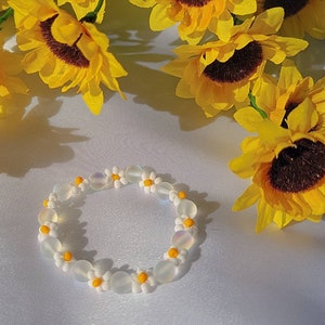 May include: A white daisy flower bead bracelet with yellow centers. The bracelet is on a white surface with yellow sunflowers in the background.