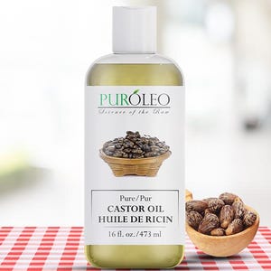 May include: A clear bottle of Puroleo castor oil with a white label featuring a basket of seeds. The label also includes the text "Science of the Raw" and "Pure Castor Oil 16 fl. oz.". A small wooden bowl filled with seeds sits to the right.