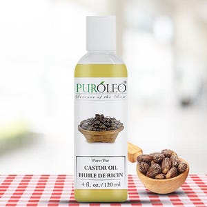May include: A clear bottle of Puroleo Castor Oil with a white cap. The label features an image of castor beans in a woven basket. The text reads "CASTOR OIL HUILE DE RICIN 4 fl. oz. / 120 ml". A small wooden bowl of castor beans sits to the right.