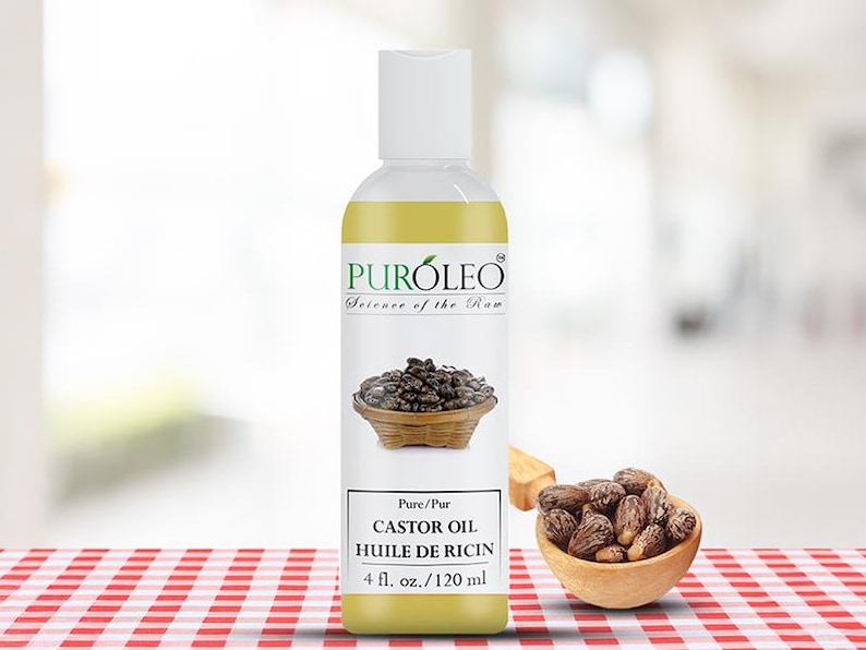 May include: A clear bottle of Puroleo Castor Oil with a white cap. The label features an image of castor beans in a woven basket. The text reads "CASTOR OIL HUILE DE RICIN 4 fl. oz. / 120 ml". A small wooden bowl of castor beans sits to the right.