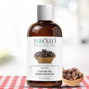 May include: A brown bottle of castor oil with a white cap and label. The label reads "PUR&Oacute;LEO Science of the Raw" and "CASTOR OIL HUILE DE RICIN 8 fl. oz./236 ml". A small wooden bowl of castor beans is next to the bottle.