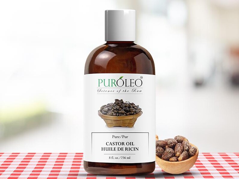 May include: A brown bottle of castor oil with a white cap and label. The label reads "PUR&Oacute;LEO Science of the Raw" and "CASTOR OIL HUILE DE RICIN 8 fl. oz./236 ml". A small wooden bowl of castor beans is next to the bottle.