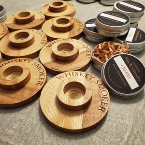 Puede incluir: Primer plano de varios soportes de madera para ahumador de whisky con el texto "Whiskey Smoker" grabado en ellos. Los soportes están hechos de madera marrón claro y tienen un pequeño agujero en el centro para una astilla de madera para ahumar. También hay varias latas metálicas pequeñas con etiquetas que dicen "956 Woodworks" y "Cherry" en ellas.