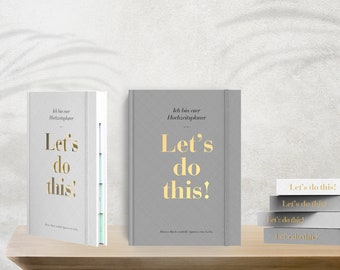May include: Two grey hardcover books with gold lettering that reads "Ich bin euer Hochzeitsplaner" and "Let's do this!" The books are on a wooden shelf in front of a grey textured background. A stack of four grey and white books with gold lettering that reads "Let's do this!" is on the shelf to the right of the two books.