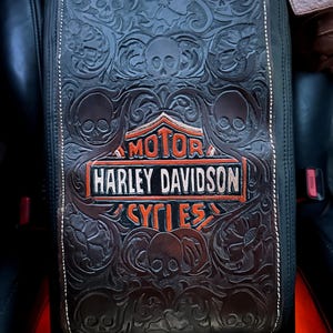 Pode incluir: Capa de console em couro marrom escuro com caveiras e padrões florais em relevo. Apresenta o logotipo da Harley Davidson em laranja e branco. Costuras brancas contornam as bordas.