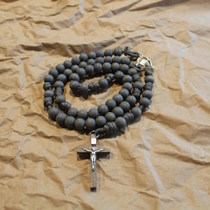 Puede incluir: Un rosario de madera gris con un crucifijo de plata. El rosario está colocado sobre un fondo de papel marrón.