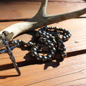 May include: A dark rosary with metallic beads and a silver crucifix. The rosary is draped over a deer antler on a wooden surface. The crucifix is detailed with a figure of Jesus.