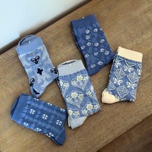Puede incluir: Cinco pares de calcetines estampados en tonos azules y crema están dispuestos sobre una superficie de madera. Los calcetines presentan varios diseños florales y geométricos. Algunos calcetines tienen un aspecto transparente y de encaje, mientras que otros tienen un tejido más sólido.