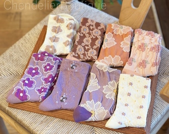 Op de afbeelding: Assortiment van acht paar sokken met bloemenmotief in verschillende kleuren, waaronder paars, bruin, crème en roze. De sokken zijn gerangschikt op een houten oppervlak.
