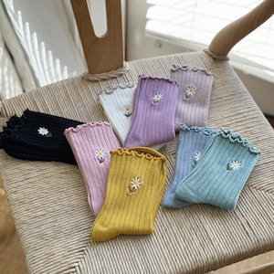 Può includere: Una collezione di calzini colorati con ricamo a margherita. I calzini sono in vari colori, tra cui nero, rosa, giallo, bianco, viola e blu. Ogni calzino ha una trama a coste e un bordo arricciato. Sono disposti su una sedia intrecciata.