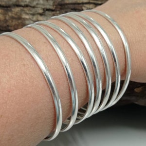 Op de afbeelding: Een set van tien zilveren armbanden gestapeld op de pols van een persoon. De armbanden zijn glad en glanzend.