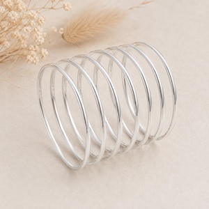 Peut inclure: Un ensemble de sept bracelets joncs fins en métal argenté poli, disposés en une formation superposée. Chaque bracelet présente une structure en fil métallique lisse et arrondi avec une finition brillante, créant un aspect circulaire uniforme.