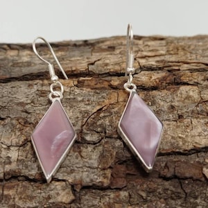 Può includere: Un paio di orecchini pendenti con una pietra a forma di diamante, viola chiaro, incastonata in una montatura in metallo color argento. Gli orecchini pendono da un semplice gancio. Lo sfondo è una superficie marrone strutturata.