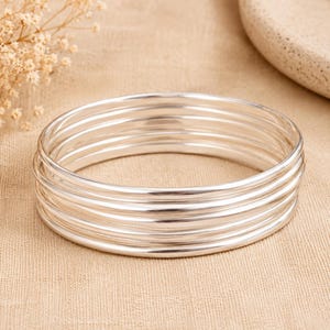 May include: A set of five stacked silver bangles featuring a smooth, polished, and reflective metallic finish. Each bangle is circular with a thin, rounded profile, creating a uniform appearance when layered together as a single jewelry piece.