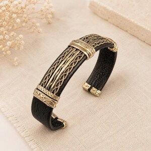 Puede incluir: Una pulsera de brazalete de cuero negro con una placa central de metal en tono dorado antiguo con detalles de alambre trenzado y tapas decorativas grabadas. El diseño abierto muestra una banda de cuero texturizado con acentos metálicos.