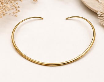 May include: A smooth, polished gold-tone metal choker necklace with a circular, open-ended design. The necklace features a thin, rounded band that curves into a wide arc, creating a simple, continuous silhouette with tapered ends.