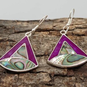 Op de afbeelding: Een paar zilveren oorbellen met een driehoekig ontwerp. De oorbellen hebben een levendige magenta rand, een centrale sectie met iriserende abalone schelp en zilveren haken. De oorbellen worden gepresenteerd op een houten oppervlak.