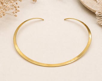 May include: A gold-toned metal choker necklace featuring a smooth, curved, open-ended band design. The piece has a polished finish that reflects light along its arc, creating a consistent metallic sheen across the entire length of the collar.