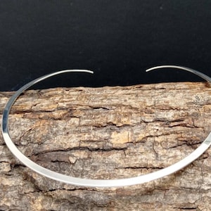 Könnte beinhalten: Eine silberne Choker-Kette aus einem flachen, breiten Metallband. Die Kette wird auf einem Stück Holz präsentiert.