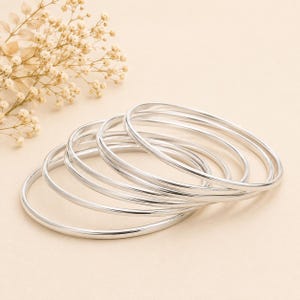 Peut inclure: Un ensemble de cinq bracelets joncs en métal argenté poli avec un profil lisse et arrondi. Les bracelets sont empilés ensemble, mettant en valeur une finition métallique réfléchissante. Ces bracelets circulaires sont fins et uniformes.