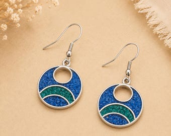 May include: A pair of silver-toned drop earrings featuring a circular frame with a cutout design. The interior of each earring is inlaid with textured blue and teal crushed stone fragments arranged in a curved, layered pattern with a hook back.