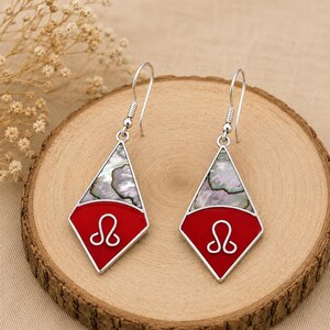 May include: A pair of silver-toned dangle earrings featuring a diamond-shaped frame. The upper section contains iridescent abalone shell inlay, while the lower section is filled with bright red enamel and a silver wire Leo zodiac symbol.
