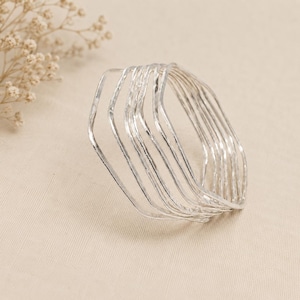 May include: A set of seven silver-colored bangles with a hammered, textured finish. Each bangle features a slightly irregular, wavy, or hexagonal shape, creating a stacked appearance. The metal surface reflects light, highlighting the hammered detail.