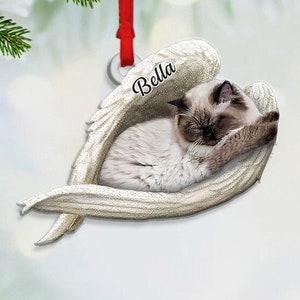 May include: A white ceramic ornament shaped like a heart with angel wings. A white cat is sleeping inside the heart. The ornament has the name "Bella" written on it.