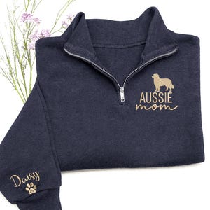 May include: Navy blue quarter-zip sweatshirt with the words "AUSSIE mom" and a gold-colored dog silhouette embroidered on the front. The sleeve has the name "Daisy" and a paw print embroidered in gold.