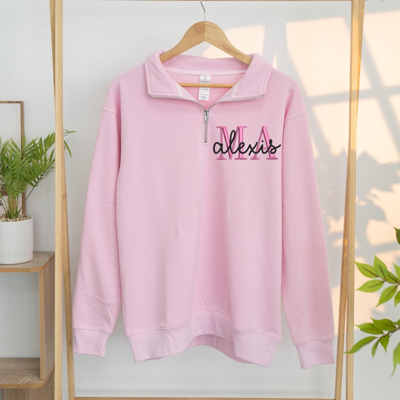 May include: A pale pink sweatshirt with a quarter-zip collar and long sleeves. The word "alexis" is embroidered in black and pink script on the front. The sweatshirt is hanging on a wooden hanger.