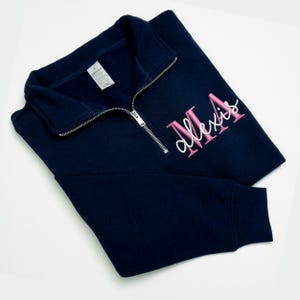 May include: A navy blue quarter-zip sweatshirt with a silver zip. The sweatshirt has the name "Alexis" embroidered in pink script with the initials "MA" in a block font. The garment is neatly folded on a white surface.