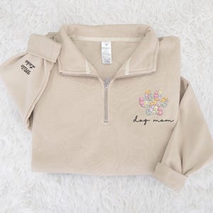May include: A beige quarter-zip sweatshirt with a floral paw print design and the words "dog mom" embroidered on the front. The sleeve cuff has the names "Milo" and "Luke" embroidered. The sweatshirt has a relaxed fit.