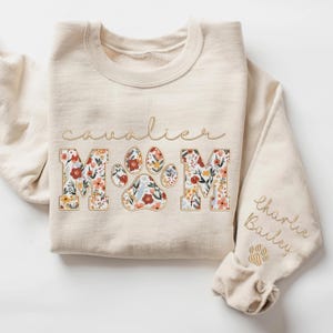 May include: A beige sweatshirt with the words "Cavalier Mom" in floral print. The word "Mom" is in large letters with a paw print in the center. The words "Charlie Bailey" are written on the sleeve with a paw print below.
