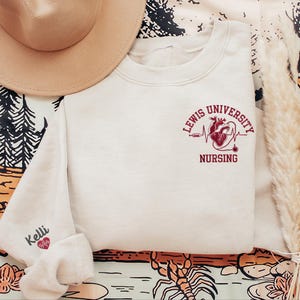 May include: A white sweatshirt with a red heart and the text "Lewis University Nursing" on the front. The sweatshirt has a small red heart with a heartbeat line on the left sleeve and the word "Kelli" above it.