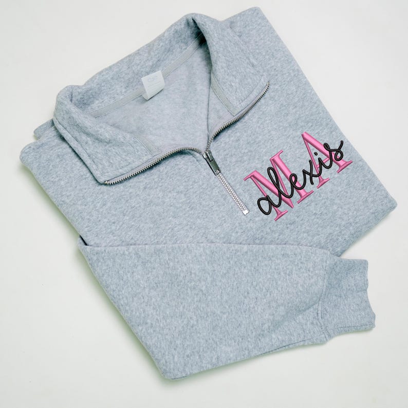 May include: A pale grey fleece pullover with a quarter-zip neckline. The word "Alexis" is embroidered in pink with a black outline on the front. The long-sleeved sweatshirt is folded neatly.