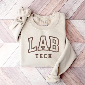 May include: A cream-colored sweatshirt with the text "LAB TECH" in brown lettering. The sweatshirt has a crew neck and long sleeves.