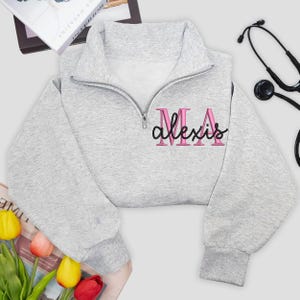 May include: A light grey cropped sweatshirt with a quarter-zip neckline. The sweatshirt features the embroidered text "MA Alexis" in pink with a black outline. A stethoscope is visible in the background.