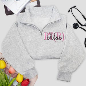 May include: A light gray cropped sweatshirt with a quarter-zip neckline. The sweatshirt features the embroidered text "PTB Chloe" in pink. A stethoscope is visible in the background. The sweatshirt is likely part of a medical professional's wardrobe.