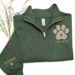 May include: A dark green quarter-zip sweatshirt with a floral paw print design and the text "DOG MOM." The sleeve features the name "COOPER" and a paw print. The sweatshirt has a silver zipper.