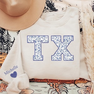 May include: A light beige sweatshirt with a blue floral print "TX" design. The sweatshirt has a small heart with the name "Michelle" embroidered on the left sleeve.