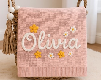 May include: A pink knitted baby blanket with the name "Olivia" embroidered in white yarn. The blanket is decorated with yellow and white embroidered flowers.